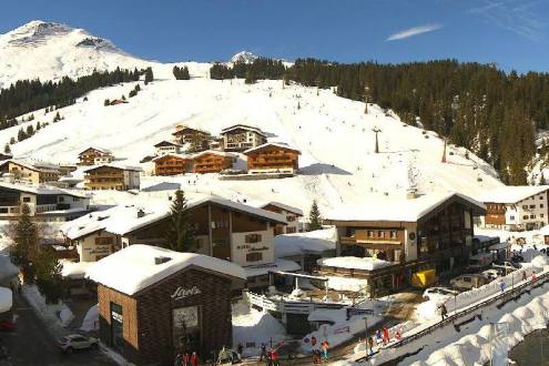 View across chalet style buildings in the centre of Lech, with blue skies above snow covered ski slopes behind the village – Weather to ski – Snow forecast, 4 February 2022