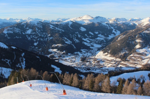 Panoramic snowy mountain view with sunny skies and Bad Kleinkirchheim, Austria, in the valley below, with a ski slope and snow cannons in the foreground – Weather to ski – Snow report, 13 January 2022