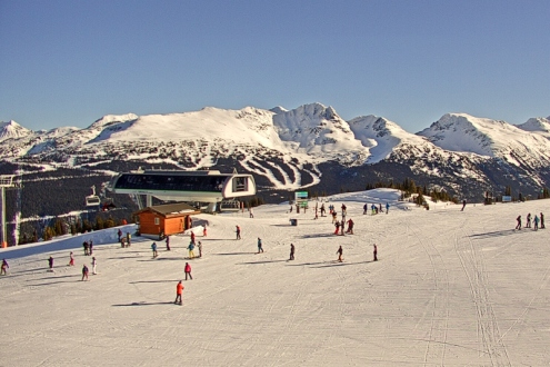Blue skies above the snowy pistes in Whistler at the summit of a chairlift with skiers in the foreground – Weather to ski – Snow report, 28 January 2022