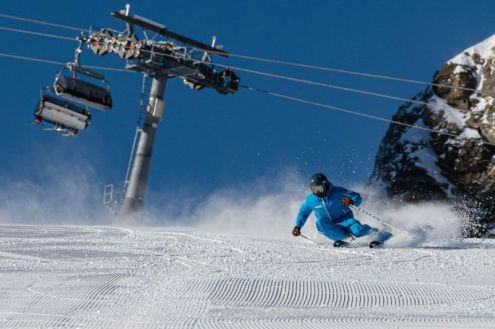 Skier in blue ski suit carving on perfectly groomed pistes kicking up snow behind them, with a chairlift behind and above, rocky terrain on the right and deep blue skies above – Weather to ski – Snow report, 3 February 2022