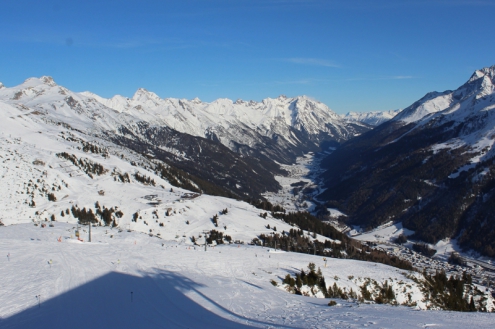 View looking down a snow-covered ski slope to the valley of St Anton, Austria, with panoramic mountain views and blue skies – Weather to ski – Half Term Snow report, 10 February 2022