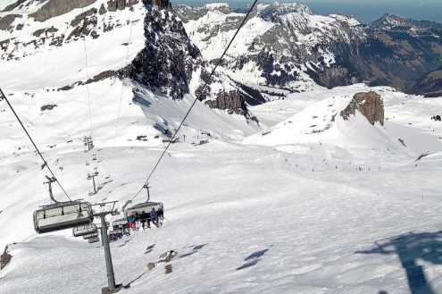 View down the line of a chairlift with seated skiers and skiers on the slopes below in Engelberg, Switzerland – Weather to ski – Snow report, 3 March 2022