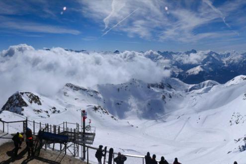 Blue skies with some low cloud above the very snowy high altitude slopes in Alpe d’Huez, France, with panoramic mountain views from a viewing platform – Weather to ski – Snow report, 15 March 2024