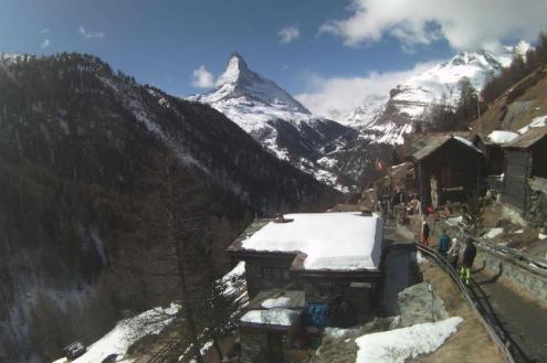 View of rustic chalet-style buildings in Findeln, with snow on the roof of a building, skiers walking carrying skis and mountain scenery in the background including the snow-covered Matterhorn mountain in Zermatt, Switzerland – Weather to ski – Snow forec