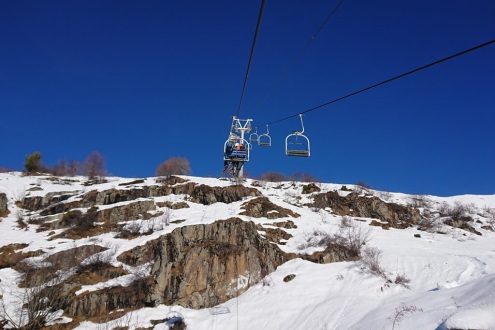 View from the Lombards chair-lift up the mountain to the Auris-en-Oisans sector of the Alpe d’Huez ski area, with a chairlift with seated skiers, snowy slopes below and blue skies above, on 21 December 2021 – Weather to ski - Our blog: Alpe d’Huez – one o