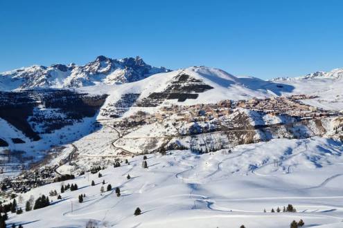 View towards Alpe d’Huez in the distance from the snowy slopes of the Auris-en-Oisans sector of the ski area, with blue skies and panoramic mountain views, on 18 December 2021 - Weather to ski – Our blog: Alpe d’Huez – one of the best all-round ski resort