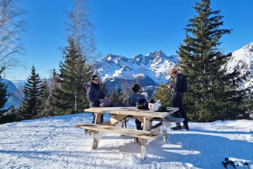 People seated and standing by picnic bench just off the Cascade run in the Montfrais sector of the Alpe d’Huez ski area, in the snow surrounded by pine trees, with blue skies above, on 19 December 2021 – Weather to ski - Our blog: Alpe d’Huez – one of the