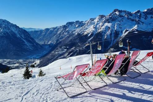 Panoramic view from the terrace of the Île d’Oz restaurant towards Oz-en-Oisans below, with blue skies above, the Alpette gondola to the right, and deckchairs with people seated in the foreground, on 19 December 2021 – Weather to ski - Our blog: Alpe d’Hu