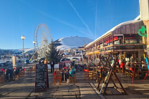 View of the Bergers commercial centre at the end of the day, with the outdoor seating of its bars and restaurants in the foreground and the Alpe d’Huez ferris wheel and ski slopes in the background, on 20 December 2021 - Weather to ski – Our blog: Alpe d’