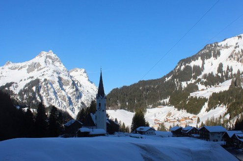 View of the village of Schröcken, Austria, with snow on the ground, view of the village church, panoramic mountain scenery behind and blue skies above – Weather to ski – Today in the Alps, 10 February 2022