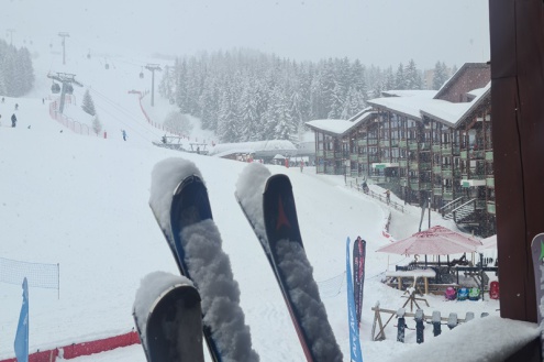 View of snow-covered skis on balcony of Tournavelles apartments at the bottom of the Front de Neige in Arc 1800, with view of pistes beyond – Weather to ski – Today in the Alps, 15 February 2022