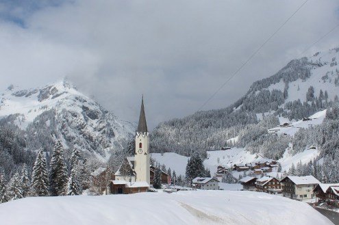 Cloudy but brightening skies and lots of snow on the ground in the resort of Schröcken, Austria, with view over the village and its church and panoramic mountain scenery beyond – Weather to ski – Today in the Alps, 3 April 2022