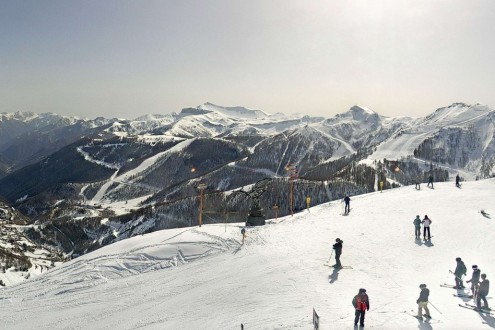 Slightly yellow skies above Auron in France, with skiers on the slopes looking at the views across the valley below – Link to Weather to ski's full "Today in the Alps" report on weather and snow in the Alps on 4 March 2026